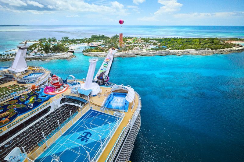 Cruise ship docked near a tropical island with water slides, a basketball court, and clear blue water.