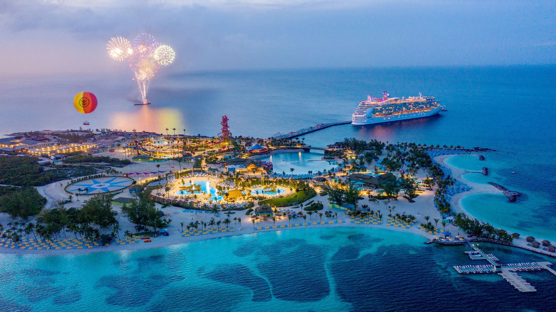 Tropical island resort at dusk with cruise ship, fireworks, hot air balloon, and lit buildings.