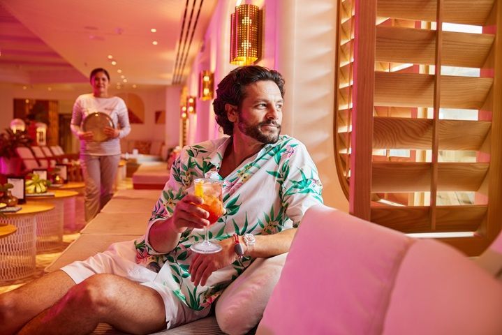 Man in tropical shirt holding a drink, looking out a window. A server walks in background. Pink and gold décor.