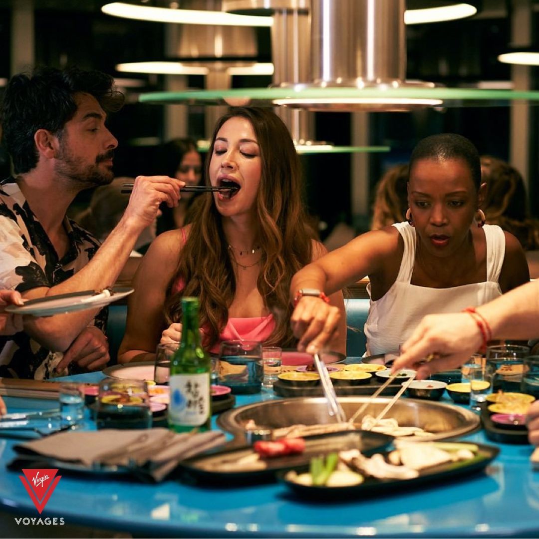 People at a Korean BBQ restaurant; a person feeds another with chopsticks. Blue table and overhead lighting.