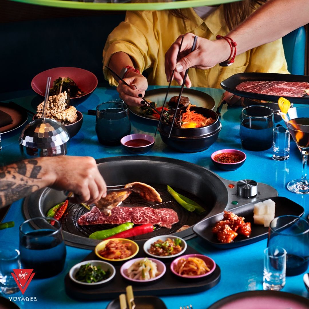 People grilling and eating Korean food at a blue table. Dishes, drinks, and red accents are visible.