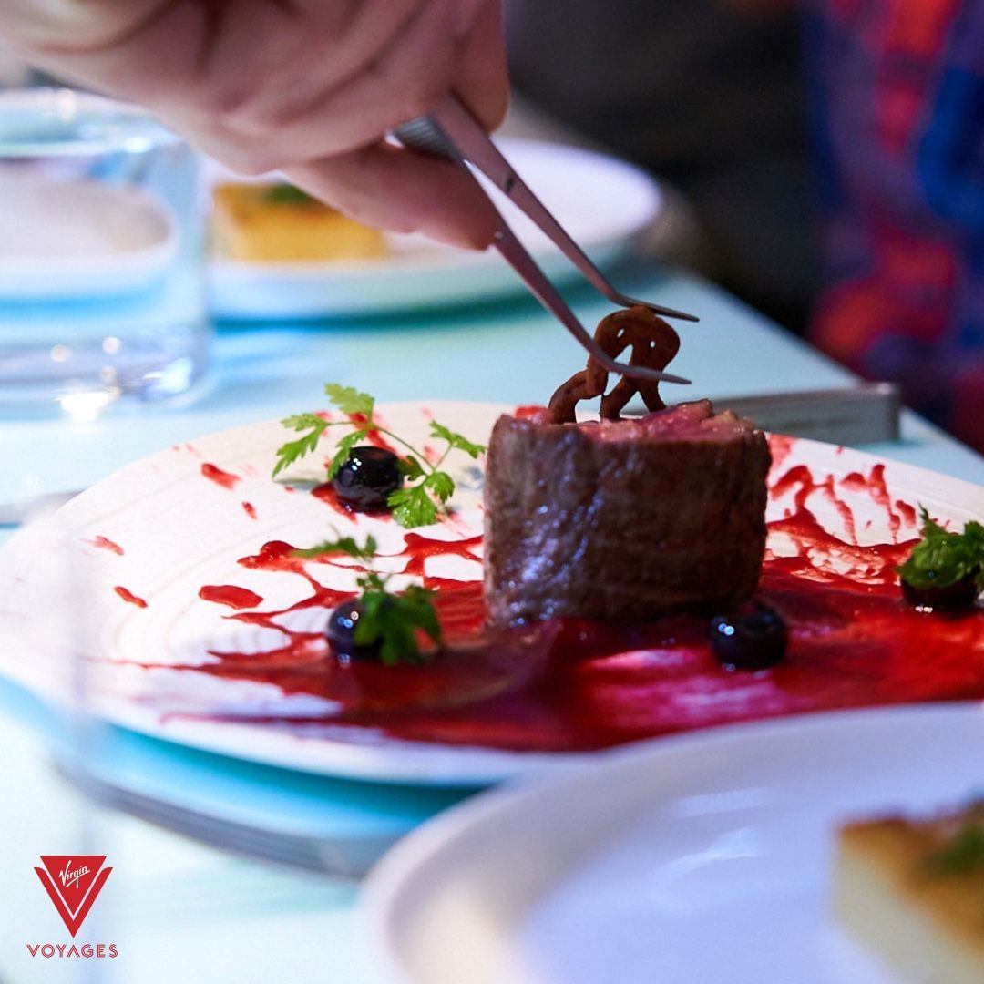 A chef using tweezers to garnish a steak with a pretzel, served on a plate with red sauce and blueberries.
