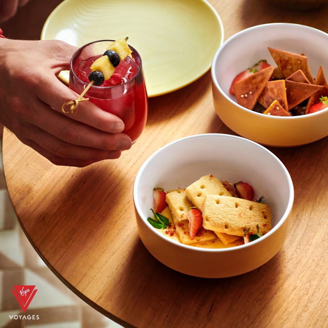 Person holding a red drink with fruit on a skewer, small bowls of snacks on a wood table.