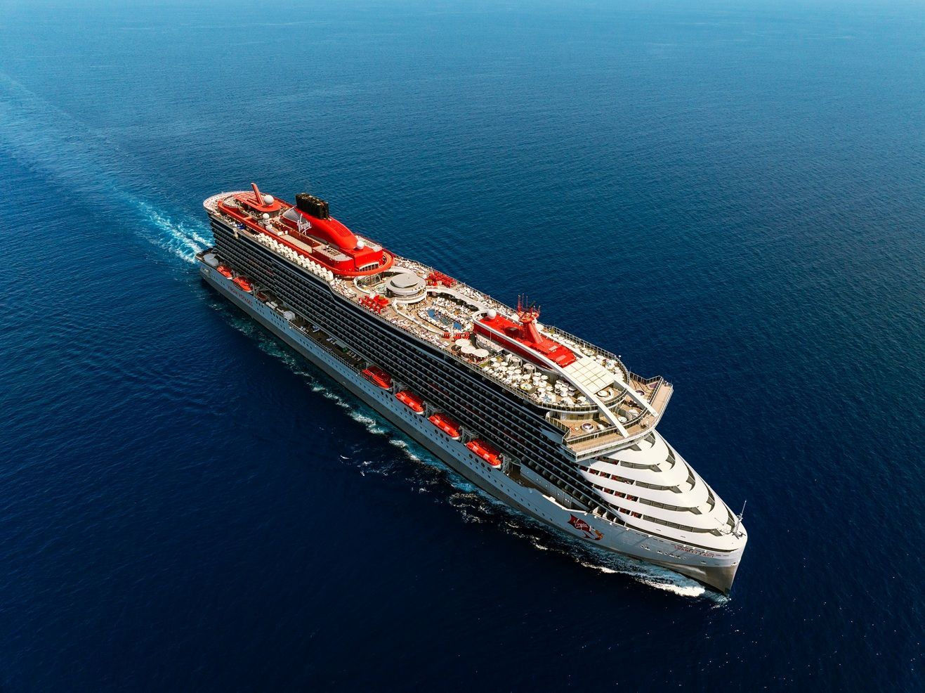 A large, modern cruise ship sails across a calm ocean under a mostly clear sky with scattered clouds. The ship features multiple decks lined with windows and balconies, red accents on the upper structures including a red funnel, and visible lifeboats along the side. A gentle wake trails behind the ship, emphasizing its motion and scale.