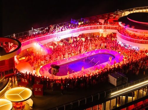 Nighttime cruise deck party with a crowd around a pool lit in red and purple lights.