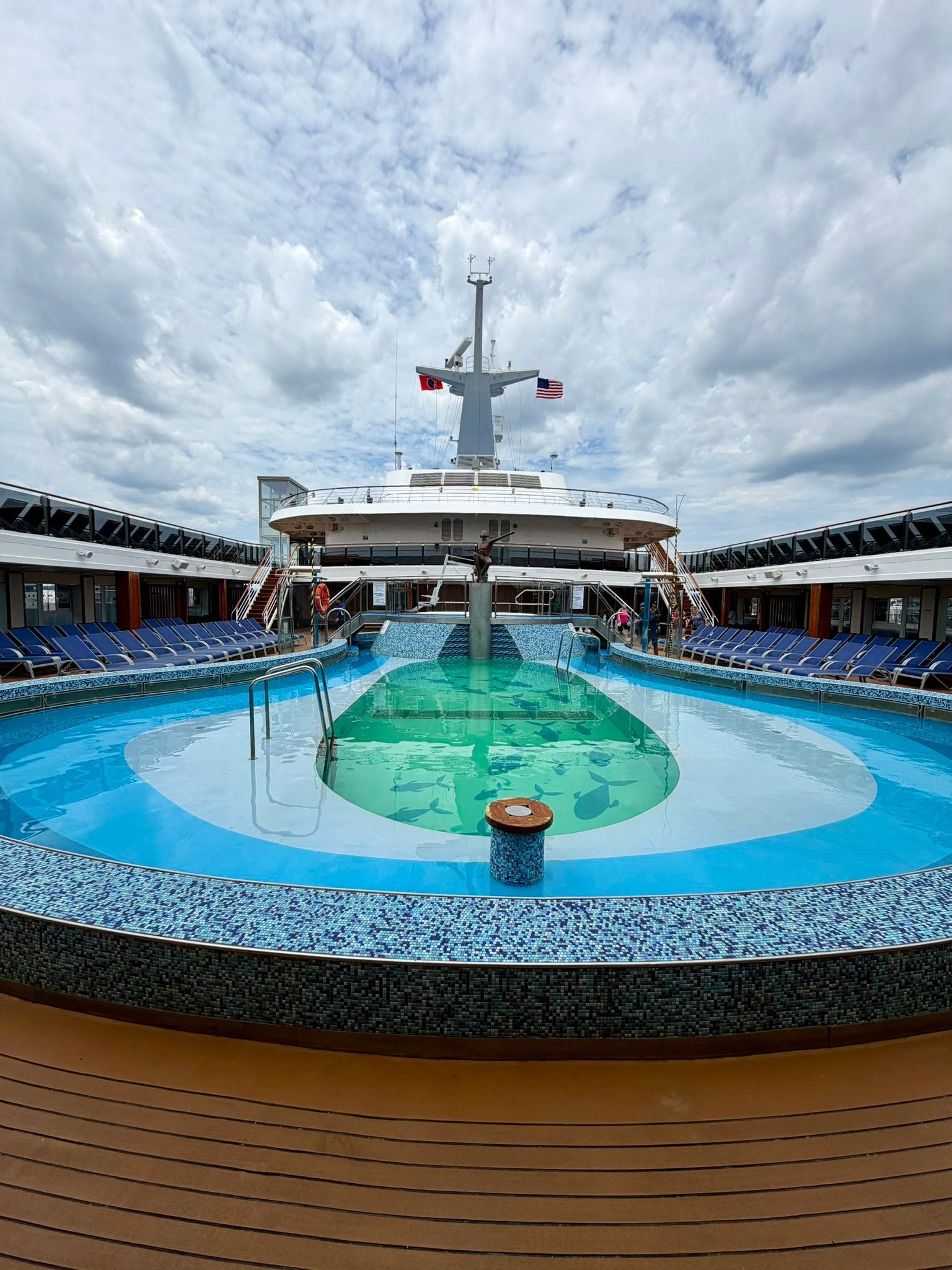Cruise ship deck with a central blue pool under a cloudy sky and rows of lounge chairs