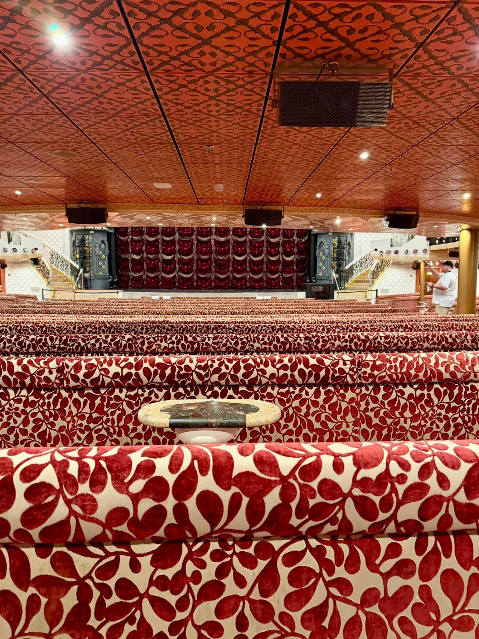 Empty banquet hall with red patterned sofas, ornate ceiling, and a stage at the far end