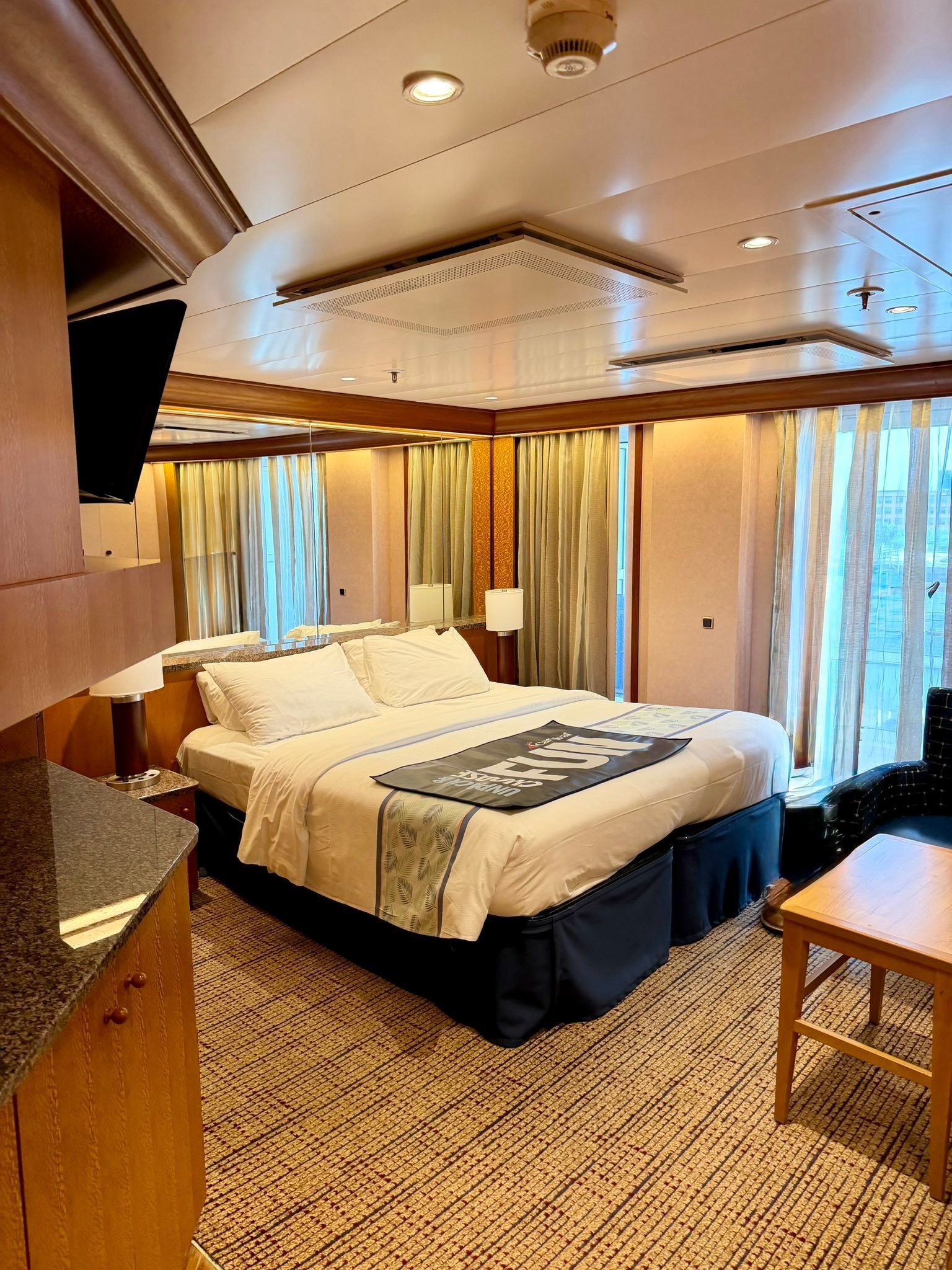 Elegant bedroom with a large bed, warm wood paneling, and floor-to-ceiling curtains on a cruise ship.