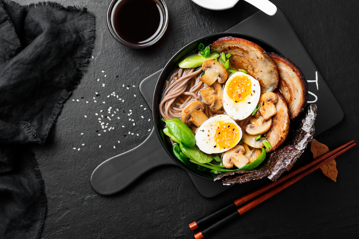 Ramen bowl with noodles, pork, mushrooms, egg, and greens; chopsticks and soy sauce on a black surface.
