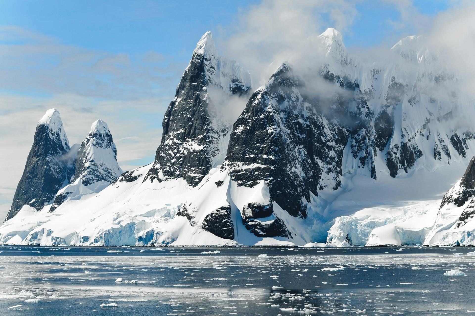 Snow-covered mountains rising from icy water, with patches of clouds and a bright blue sky.