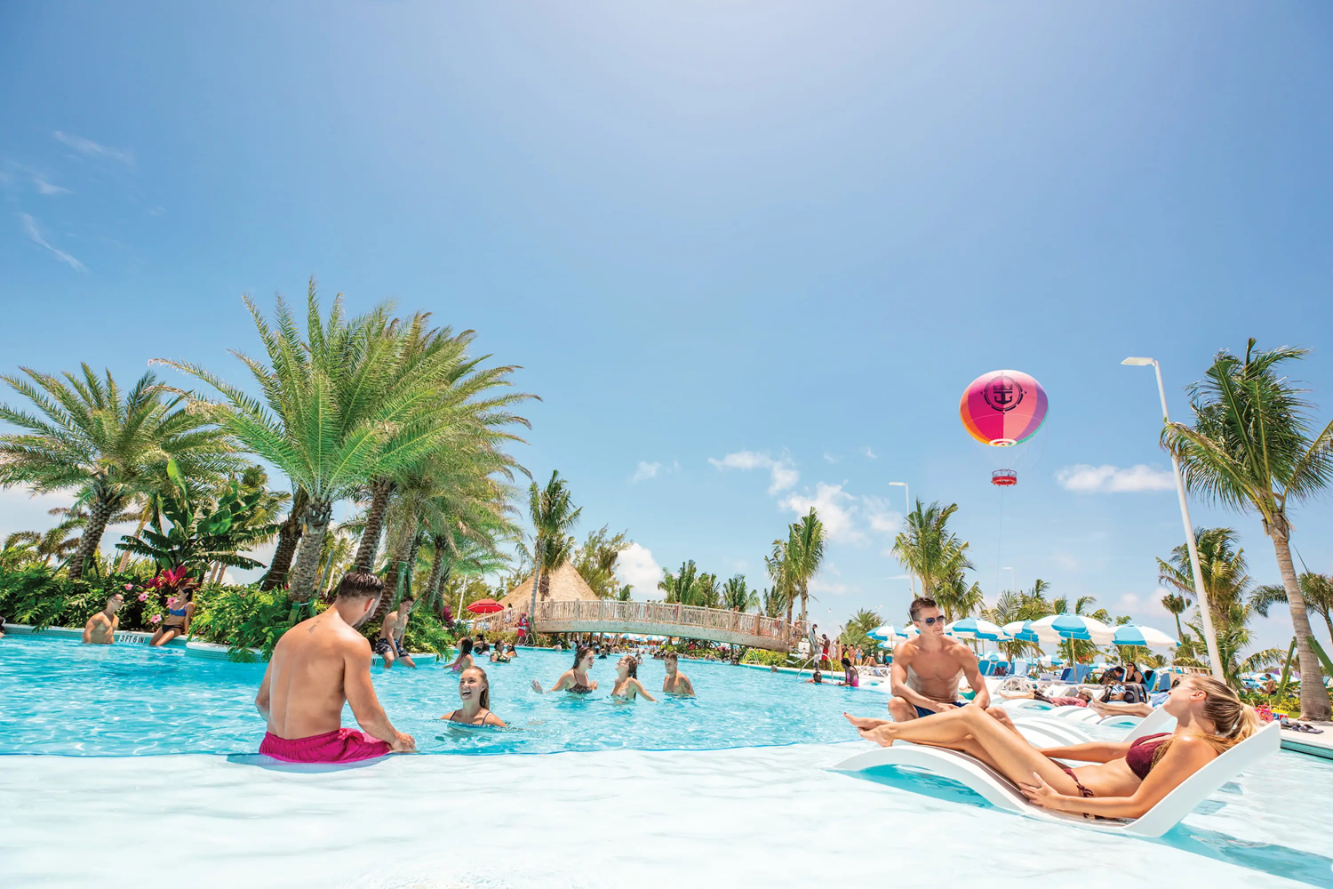 People relaxing and swimming in a tropical pool with palm trees, a hot air balloon, and a bridge.