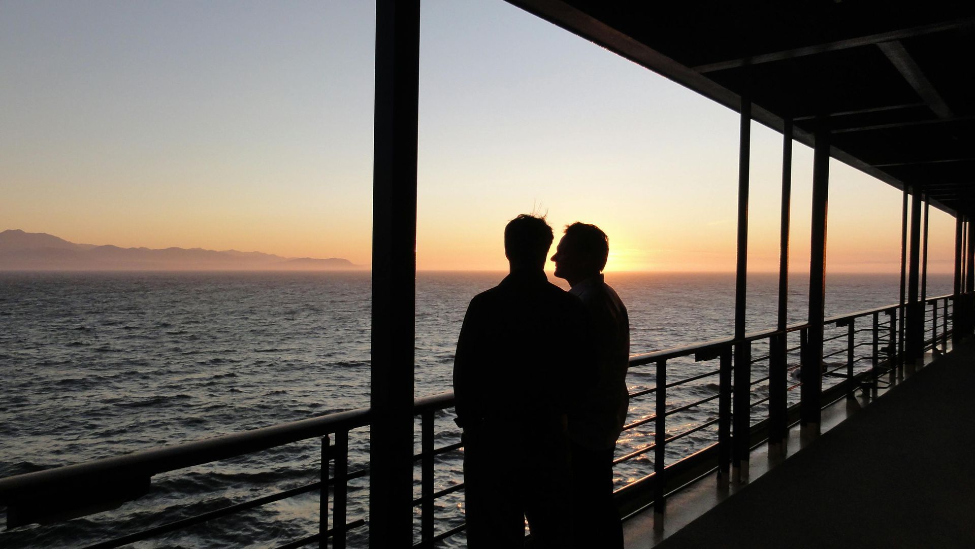 Two silhouetted people on a ship deck watch the sunset over the ocean.