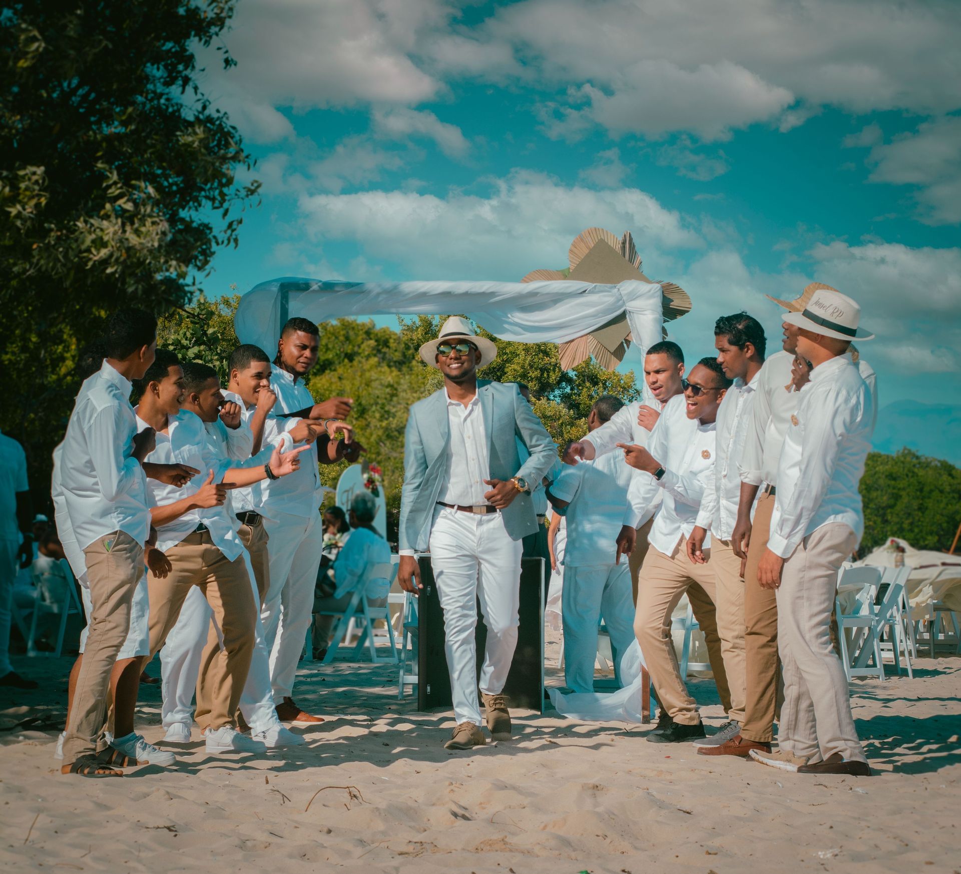 Group of people in white outfits celebrating on a beach under a blue sky, dancing by a decorated arch.
