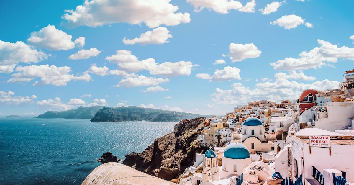 White buildings with blue domes on a cliff overlooking the Aegean Sea under a blue sky with clouds.