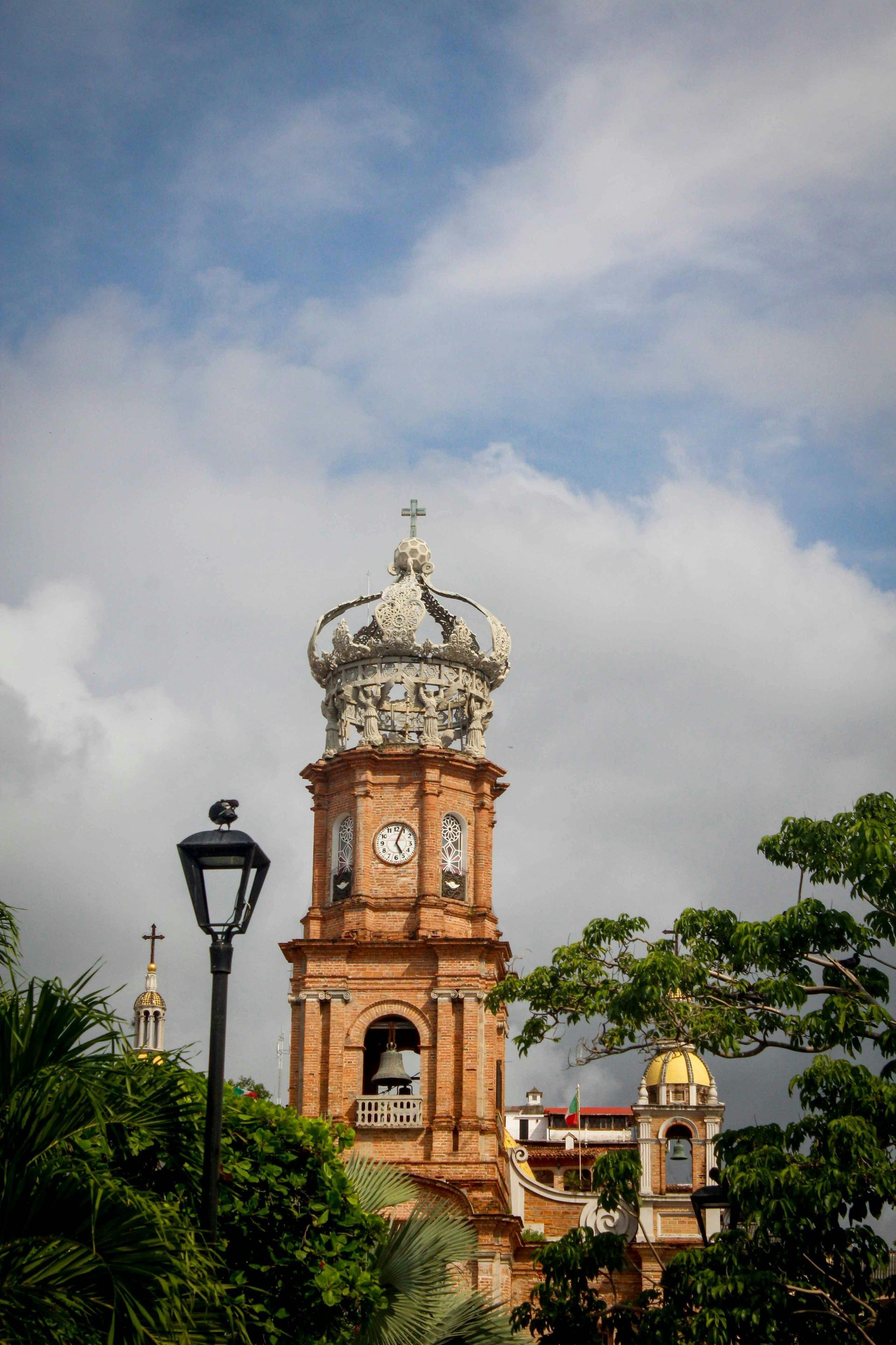 Historic church tower with ornate dome and cross against a cloudy blue sky, framed by greenery and street lamps