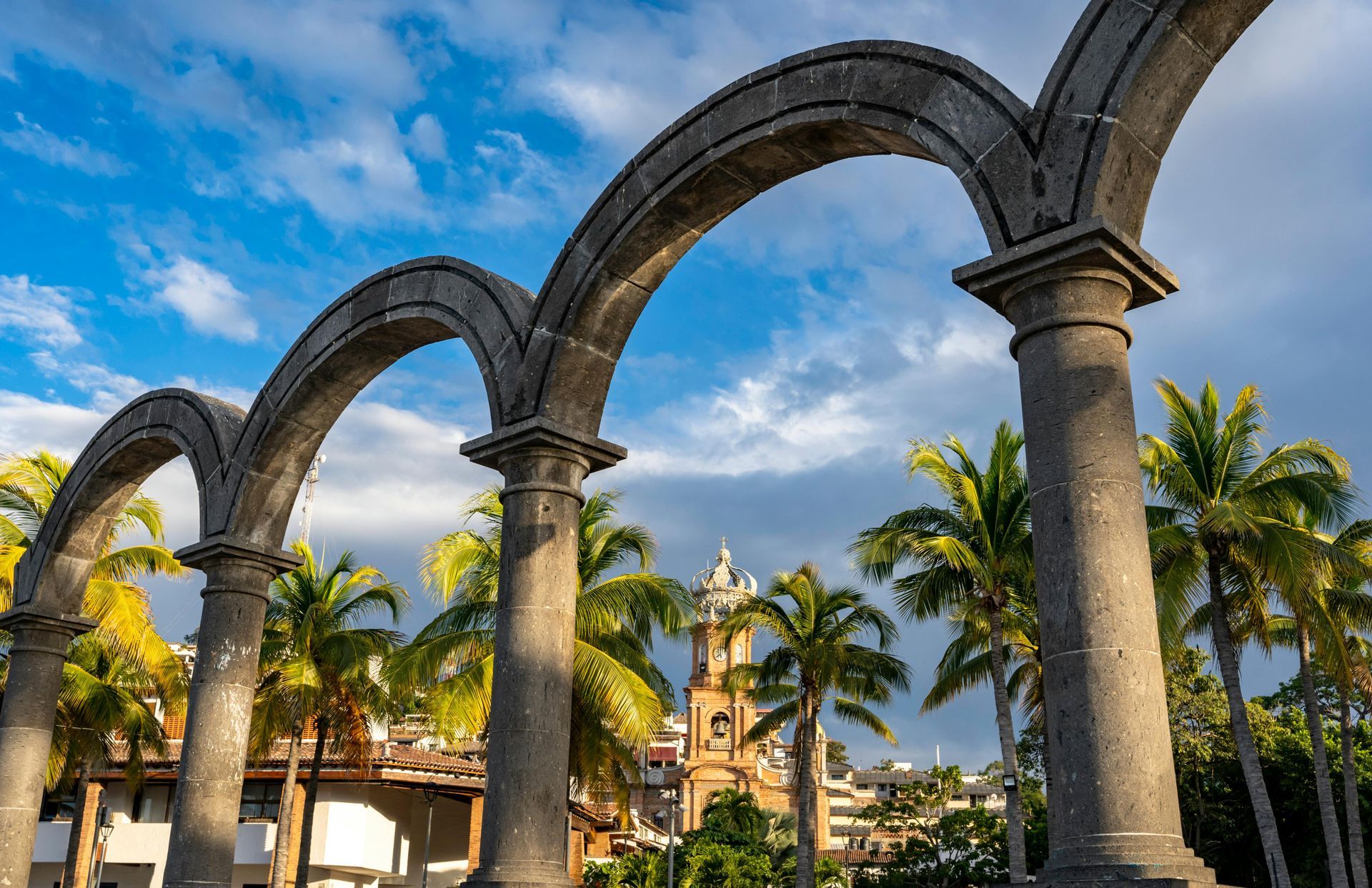 Stone arches and columns in a sunny plaza with palm trees and a historic building under a blue sky