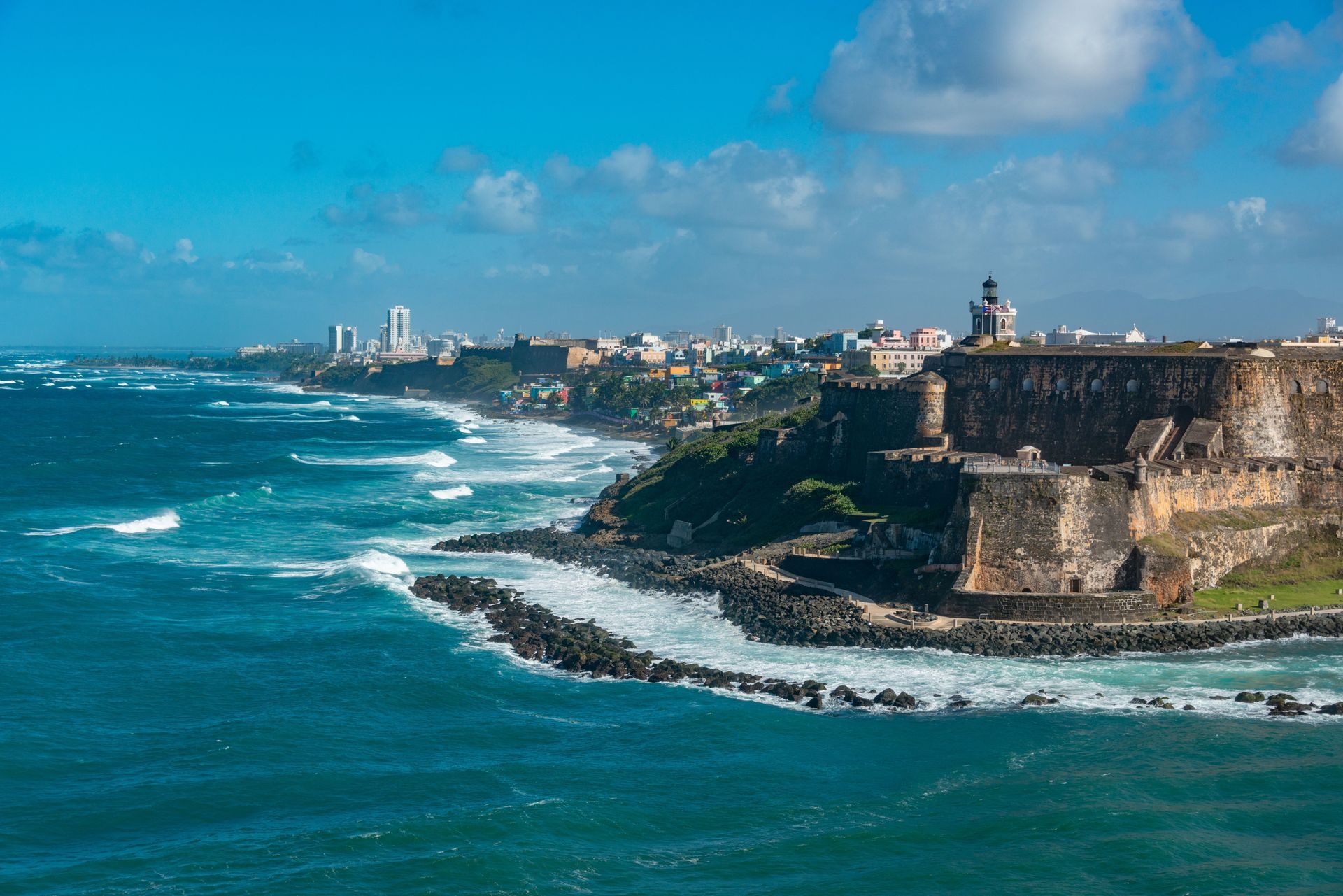 Coastal fortress overlooking a blue ocean with white surf and a distant city skyline under a clear sky