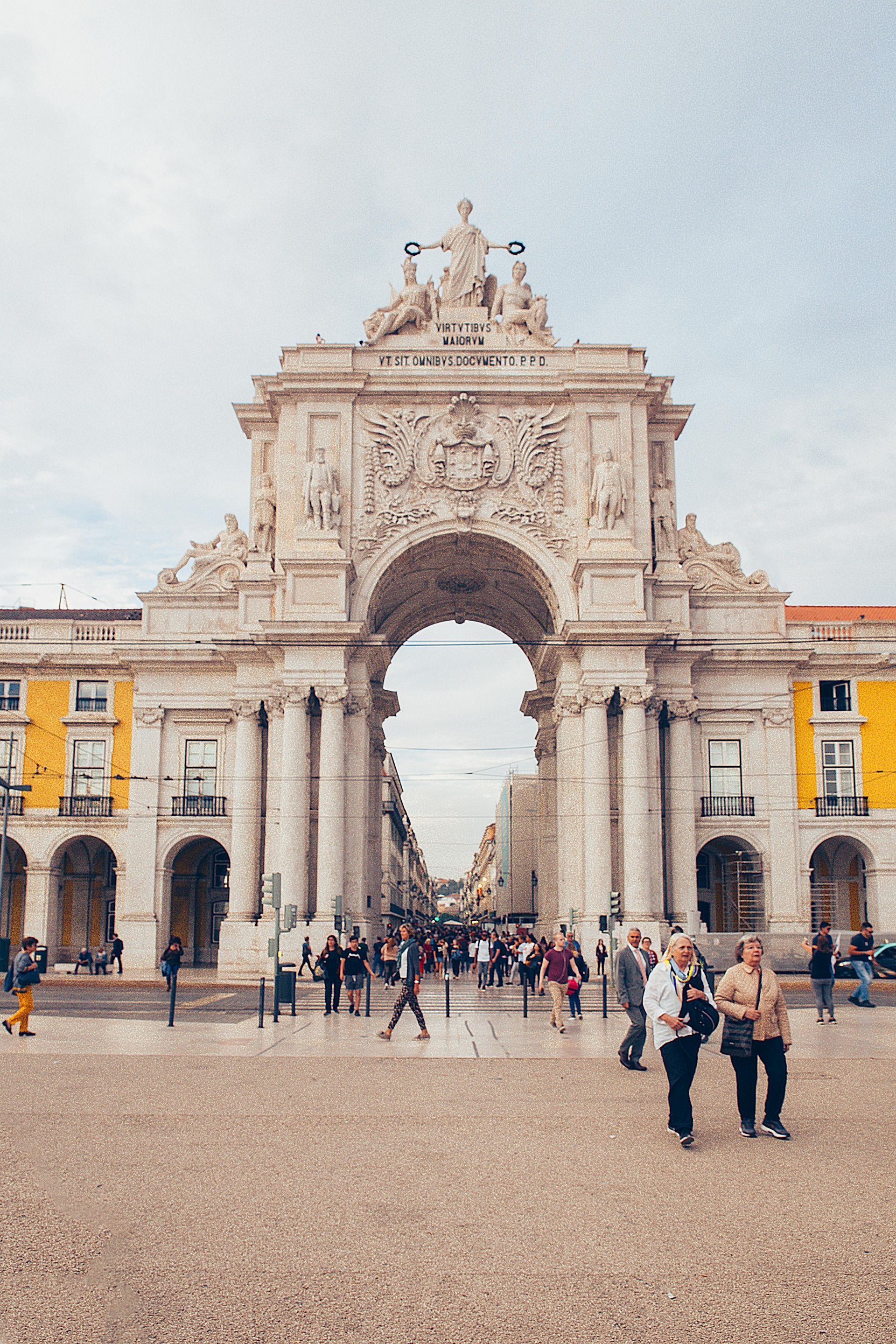 Triumphal Arch in Lisbon with people walking in a plaza. Beige, white and yellow buildings and sky.