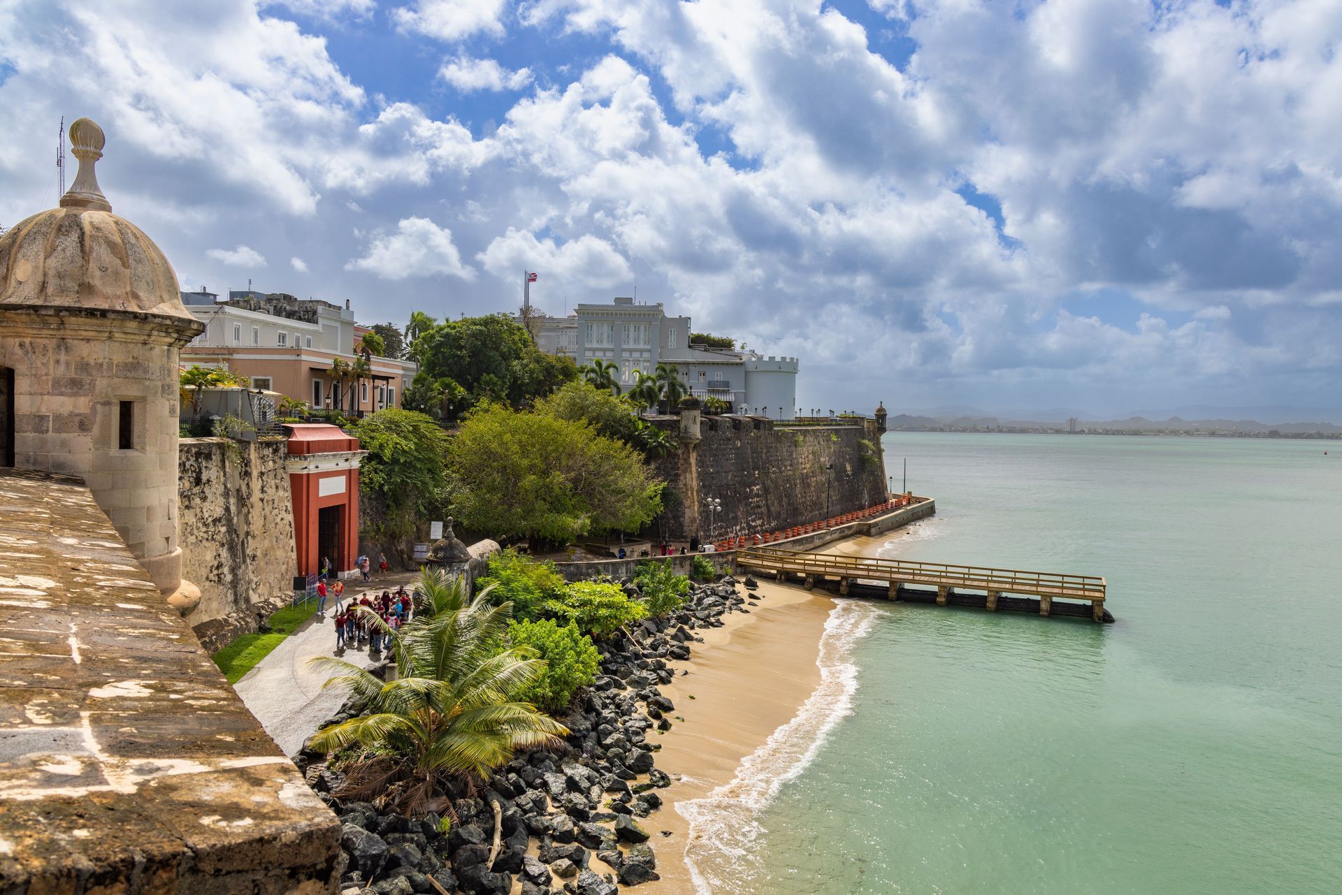 Seaside stone fort and walkway beside calm green water under cloudy sky