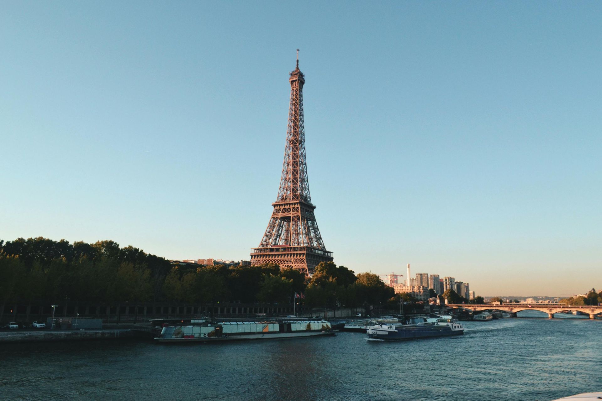 Eiffel Tower over the Seine River in Paris; boats and trees along the water with clear blue sky.