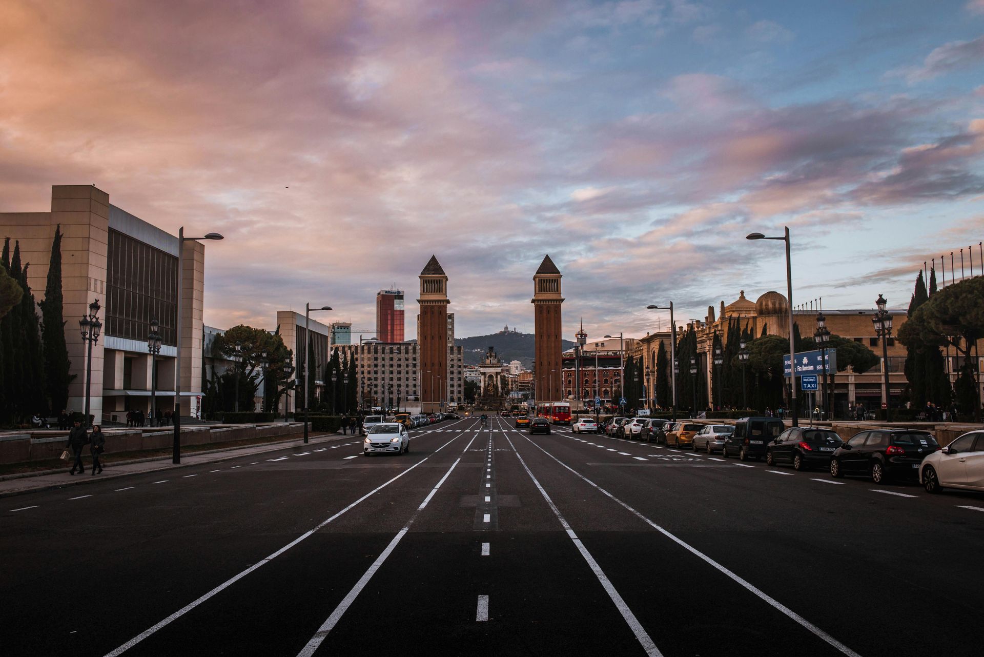 Wide street in Barcelona, Spain, leads to two tall towers. Cars parked on the sides, trees line the street, sunset sky.