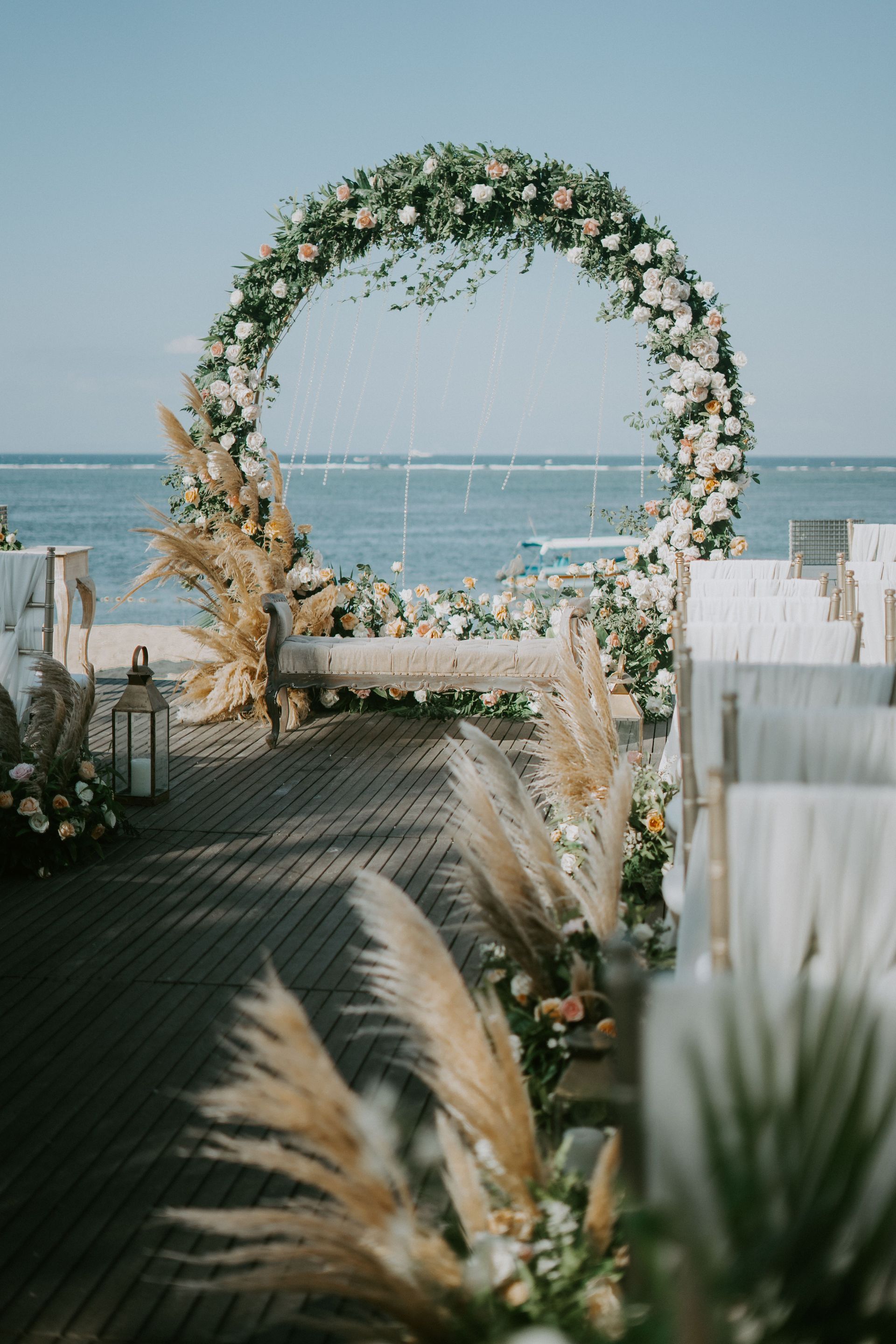 Floral wedding arch on a beach aisle with ocean backdrop and white chairs