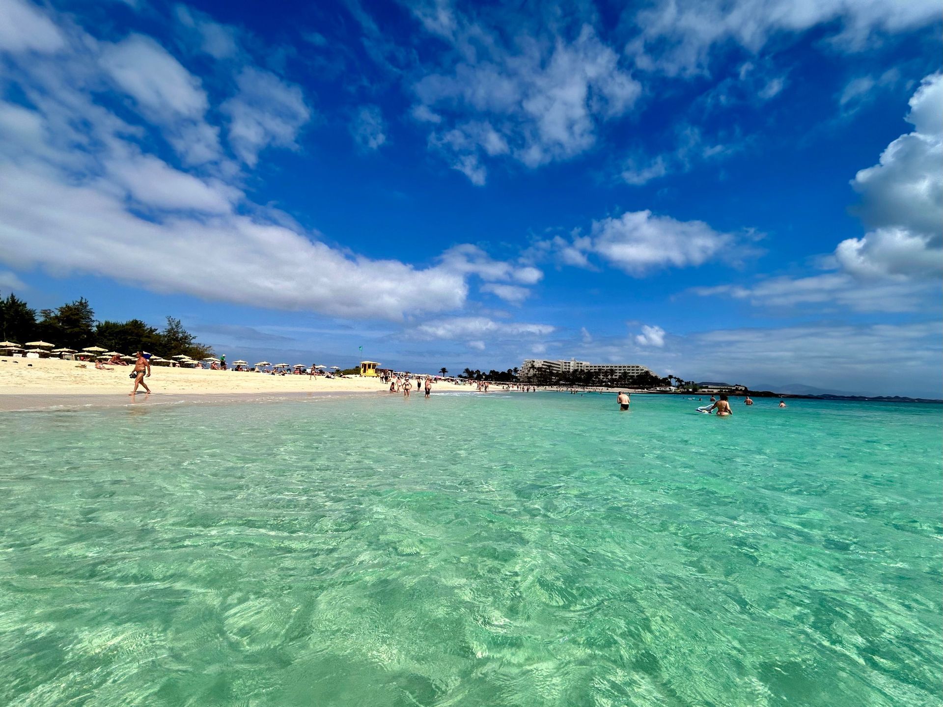 Turquoise beach water with swimmers, sandy shore, and a bright blue sky with clouds