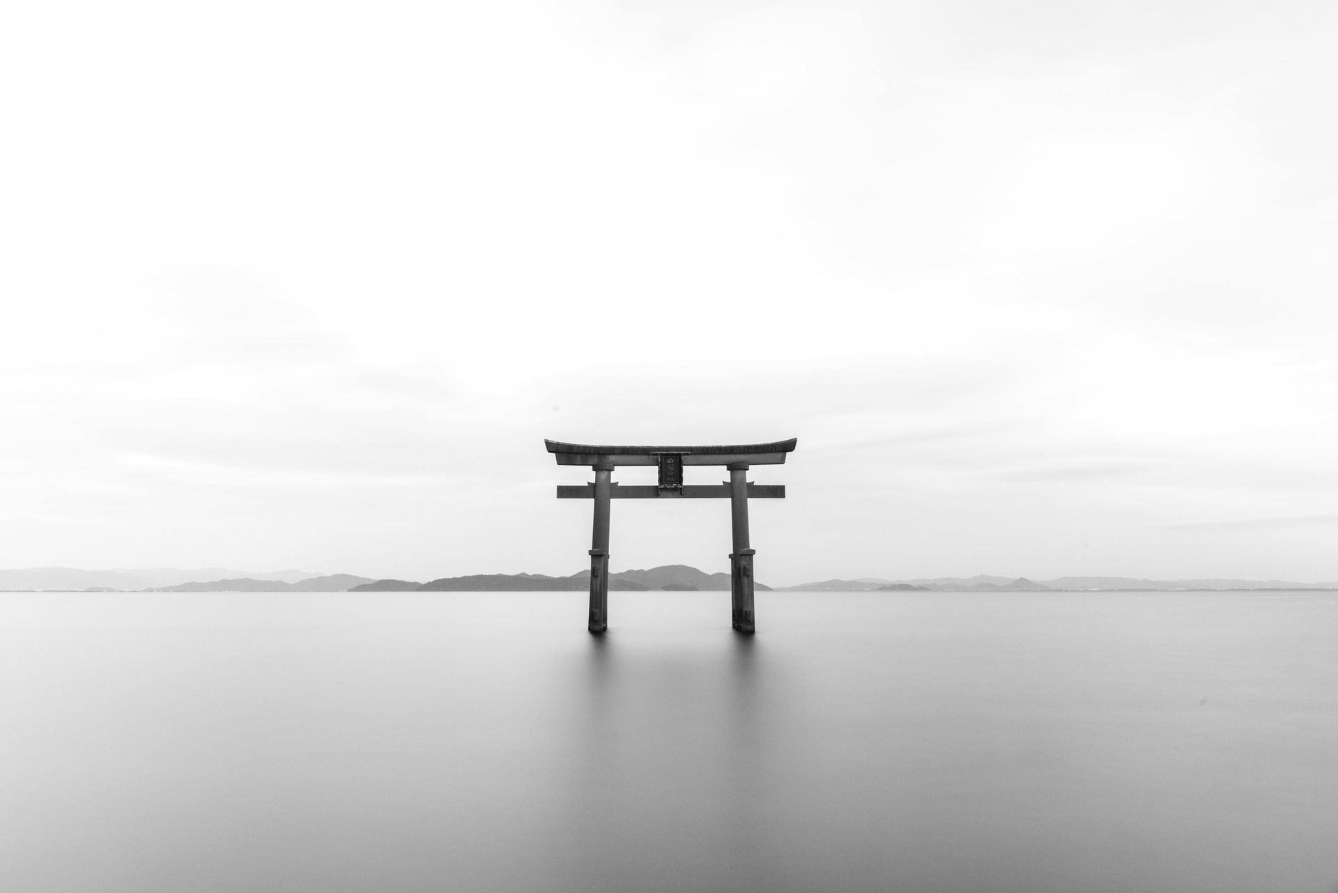 Black and white photo of a torii gate standing in calm water, with a blurred horizon in the background.
