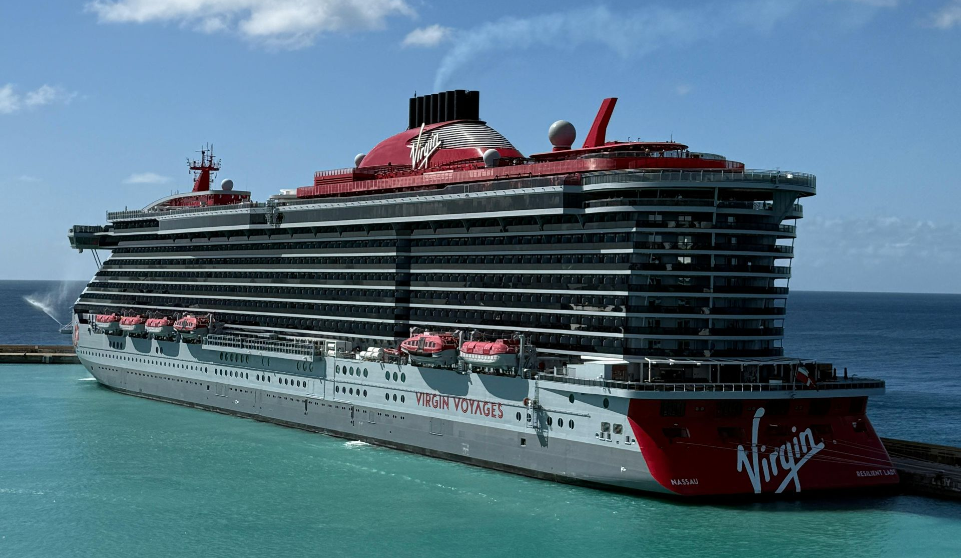 A large Virgin cruise ship with red and grey exterior on a sunny day near the ocean.