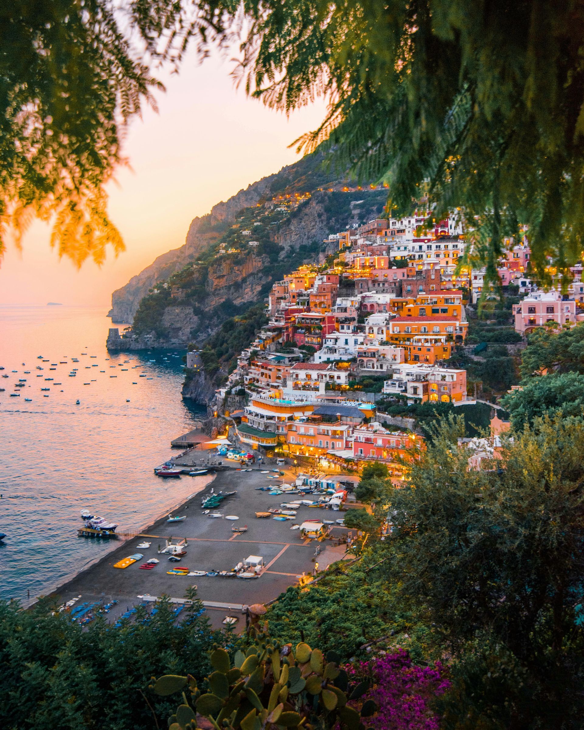 Coastal Italian town nestled into a cliff, illuminated at sunset with boats in the sea and foliage framing the view.