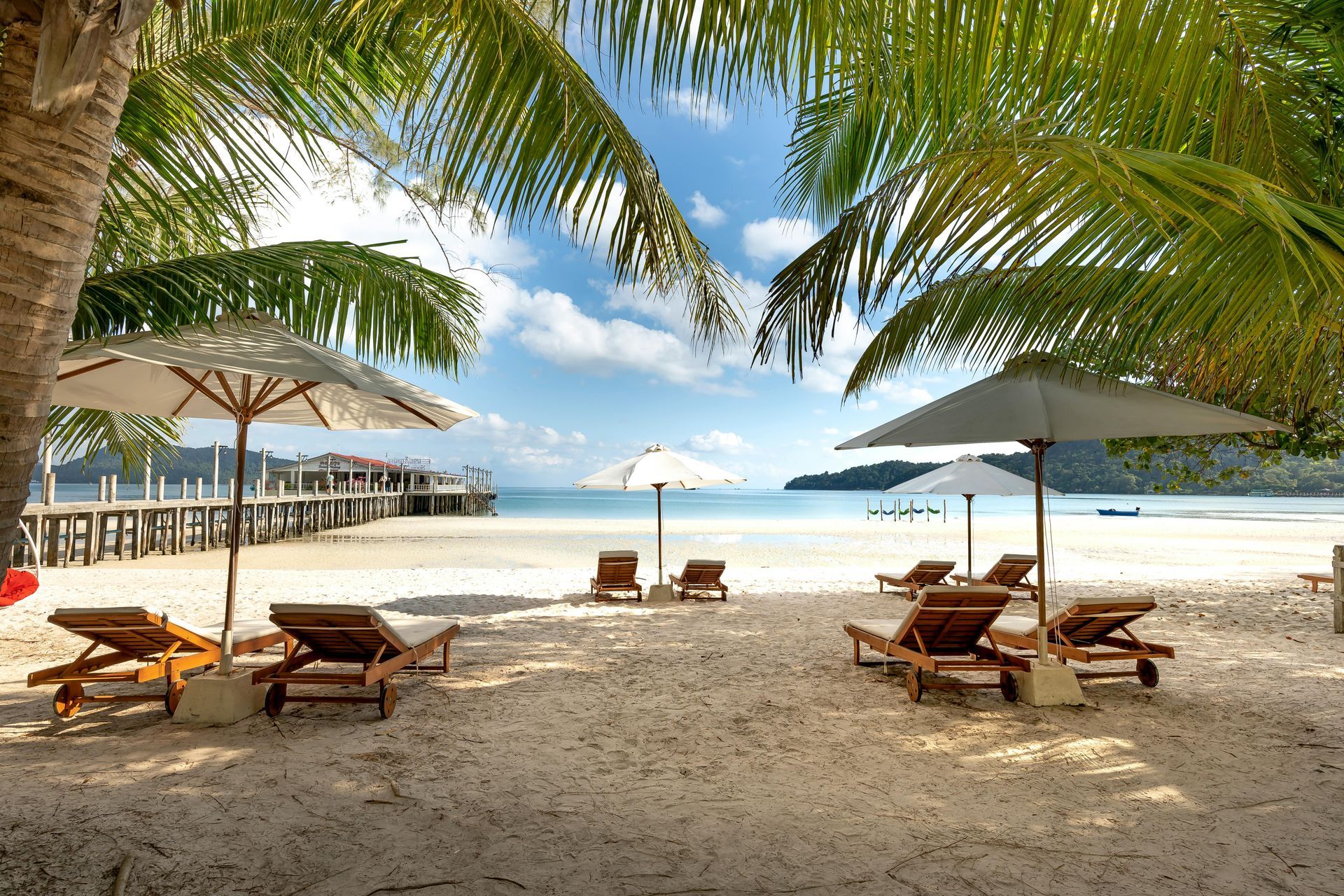 Beach scene with lounge chairs, umbrellas, palm trees, and a pier on a sunny day.