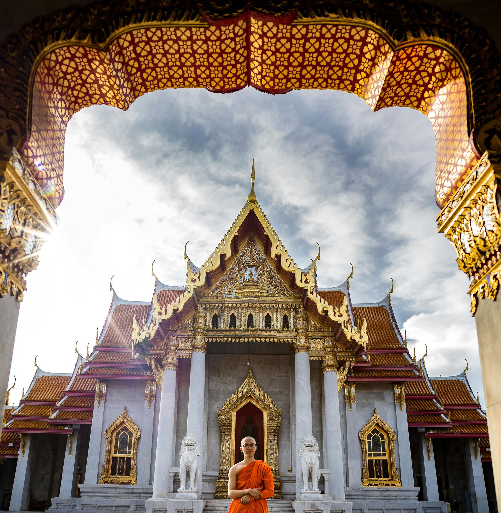Monk in orange robes stands before ornate temple, golden details, bright sky.