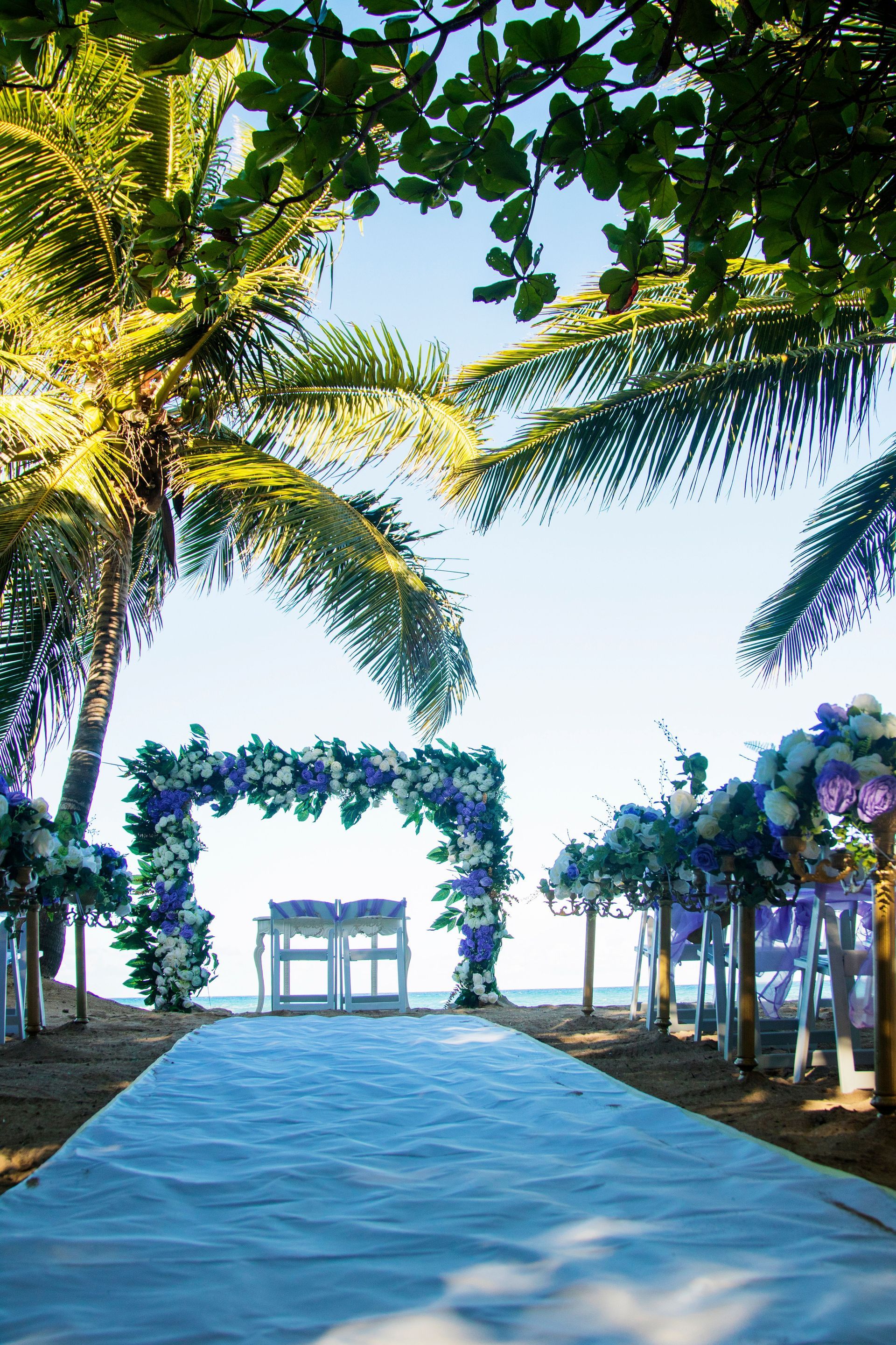 Palm-lined sandy aisle leading to a floral arch and oceanfront gazebo on a sunny beach