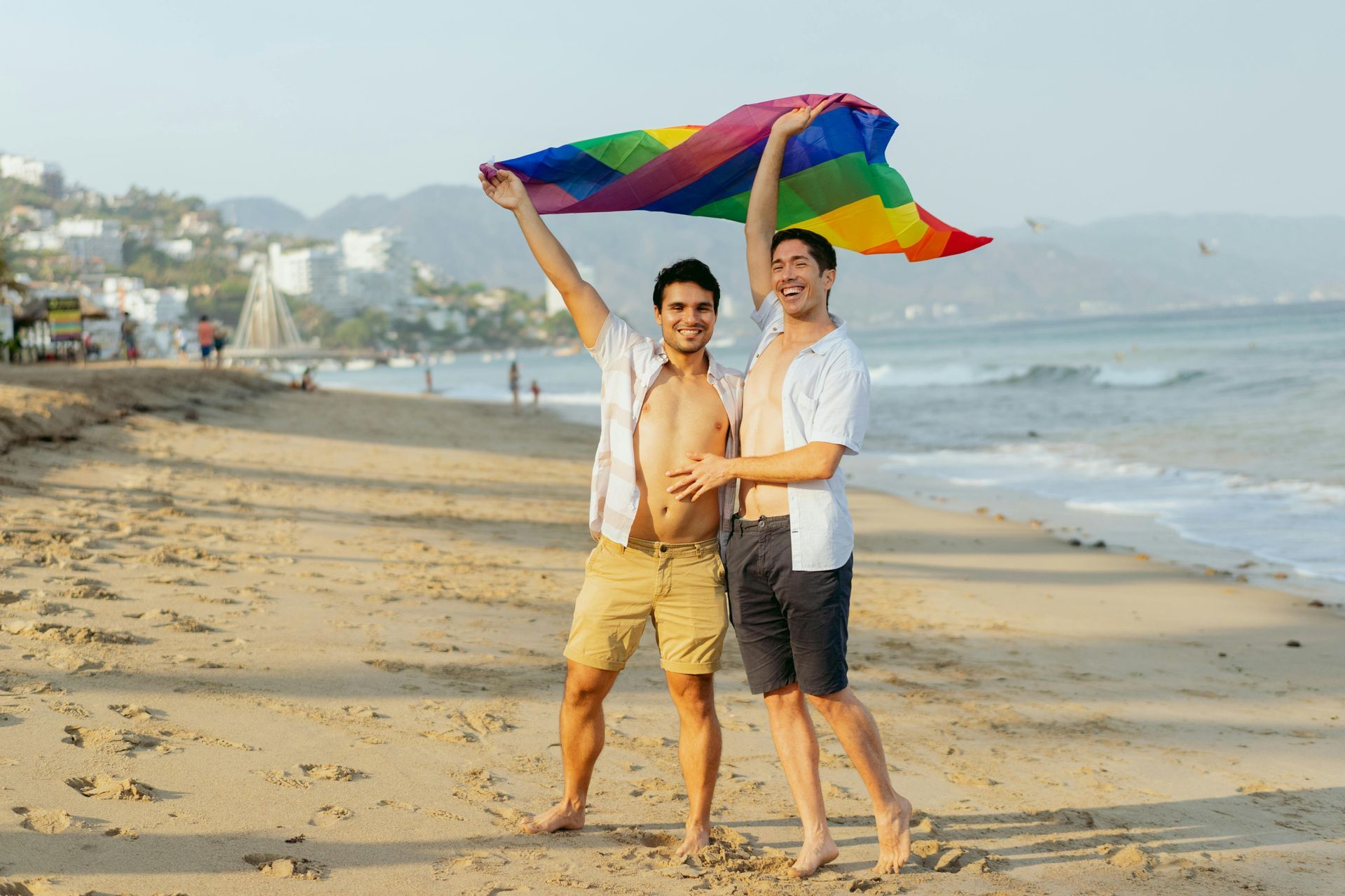 Two people on a beach hold a rainbow flag above them. They both smile. Sunny day.