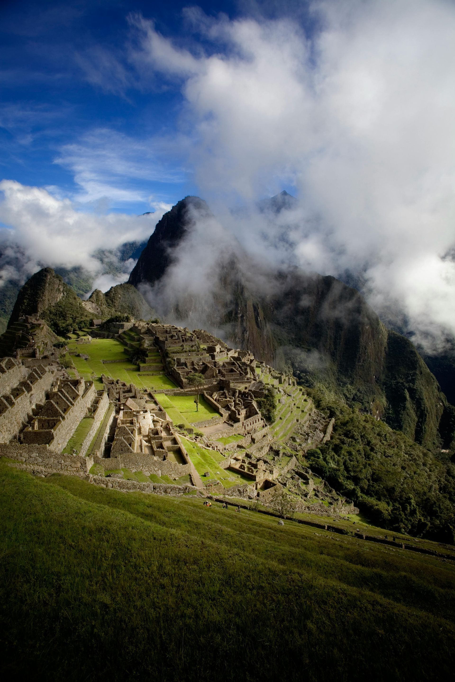 Machu Picchu ruins on a green hillside, partly obscured by clouds, under a blue sky.