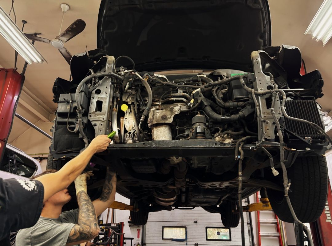 Car lifted on a garage hoist, exposing the undercarriage while a mechanic points at the engine bay. | Suburban Wrench