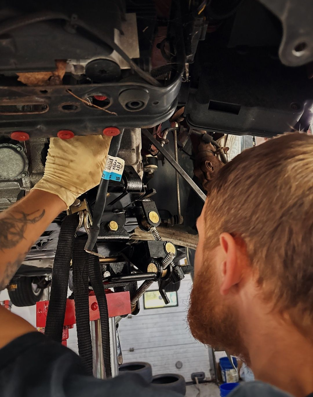 Mechanic working under a vehicle, inspecting exposed parts with a glove and tool. | Suburban Wrench