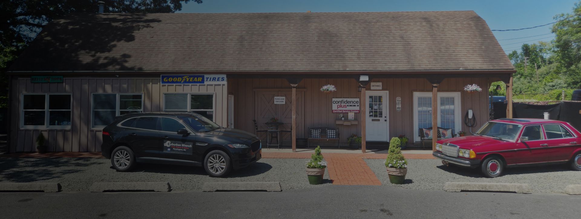 Small roadside shop with parked black and red cars under a shingled roof on a sunny day | Suburban Wrench