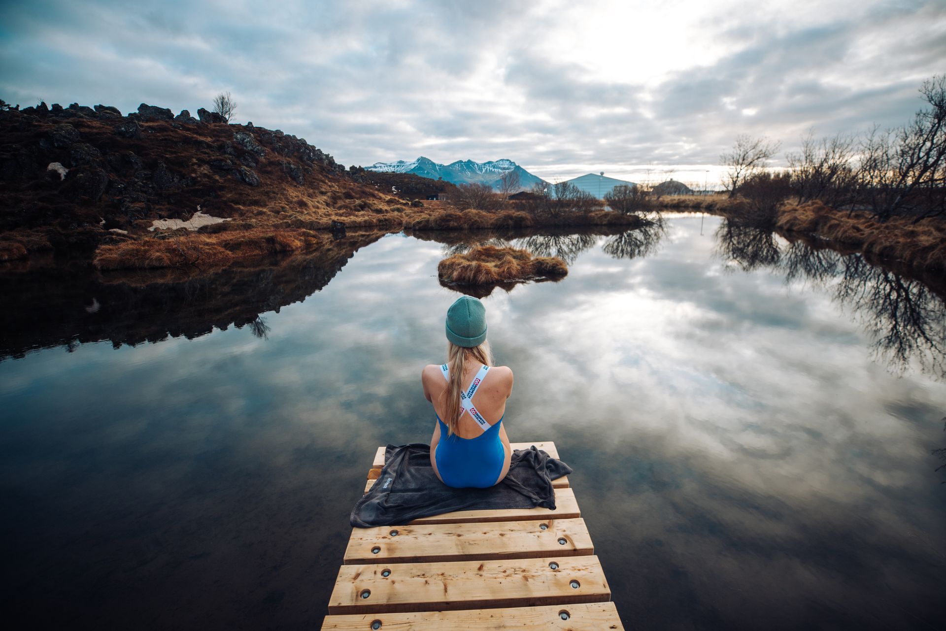 A person in a blue swimsuit and beanie sits on a wooden dock overlooking a calm, reflective lake under a cloudy sky.