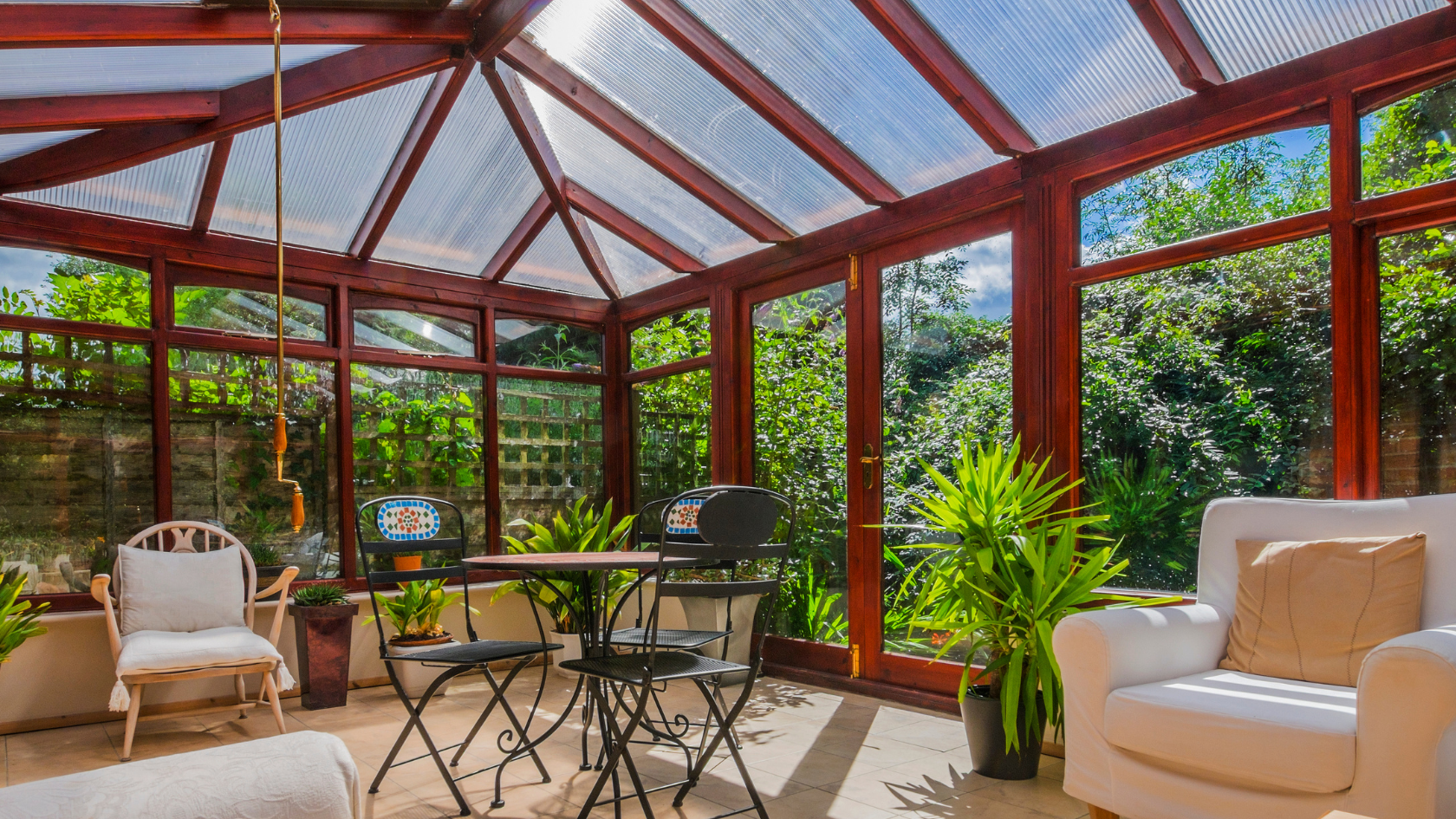 Sunroom with wooden frame, glass windows, plants, table, chairs, and white armchairs.