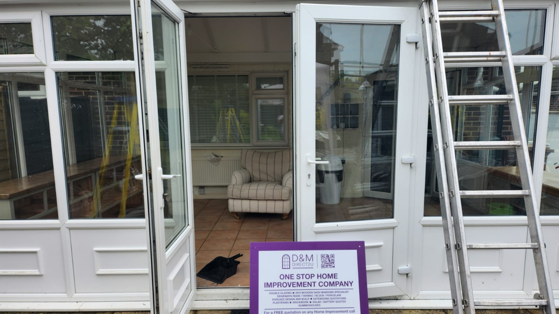 White-framed sunroom with open doors. Ladder propped up, chair inside. Sign on the ground.