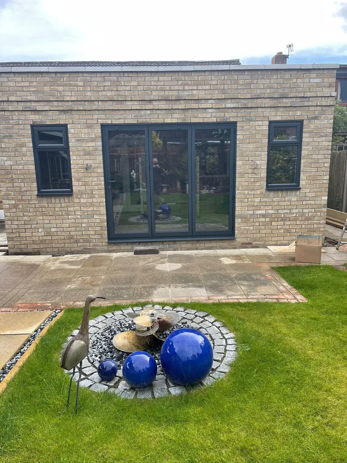 Stone building with dark windows and doors, patio, and garden with blue spheres and metal bird sculpture.