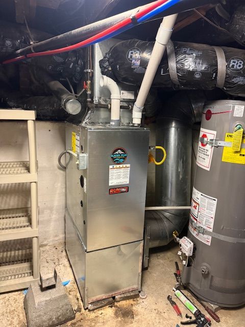 Furnace and water heater in a narrow basement utility room with exposed pipes and ductwork.