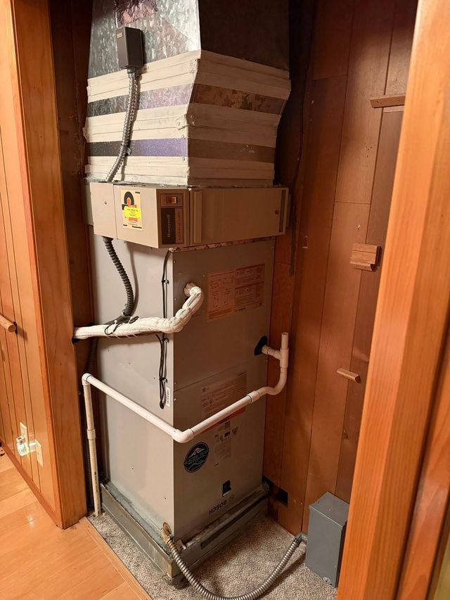 Large HVAC unit in a narrow wooden utility closet with exposed pipes and ducts.