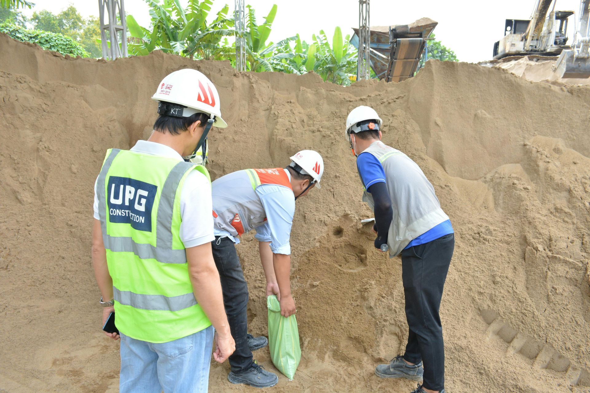 Three workers in safety gear inspecting and handling sand at a construction site