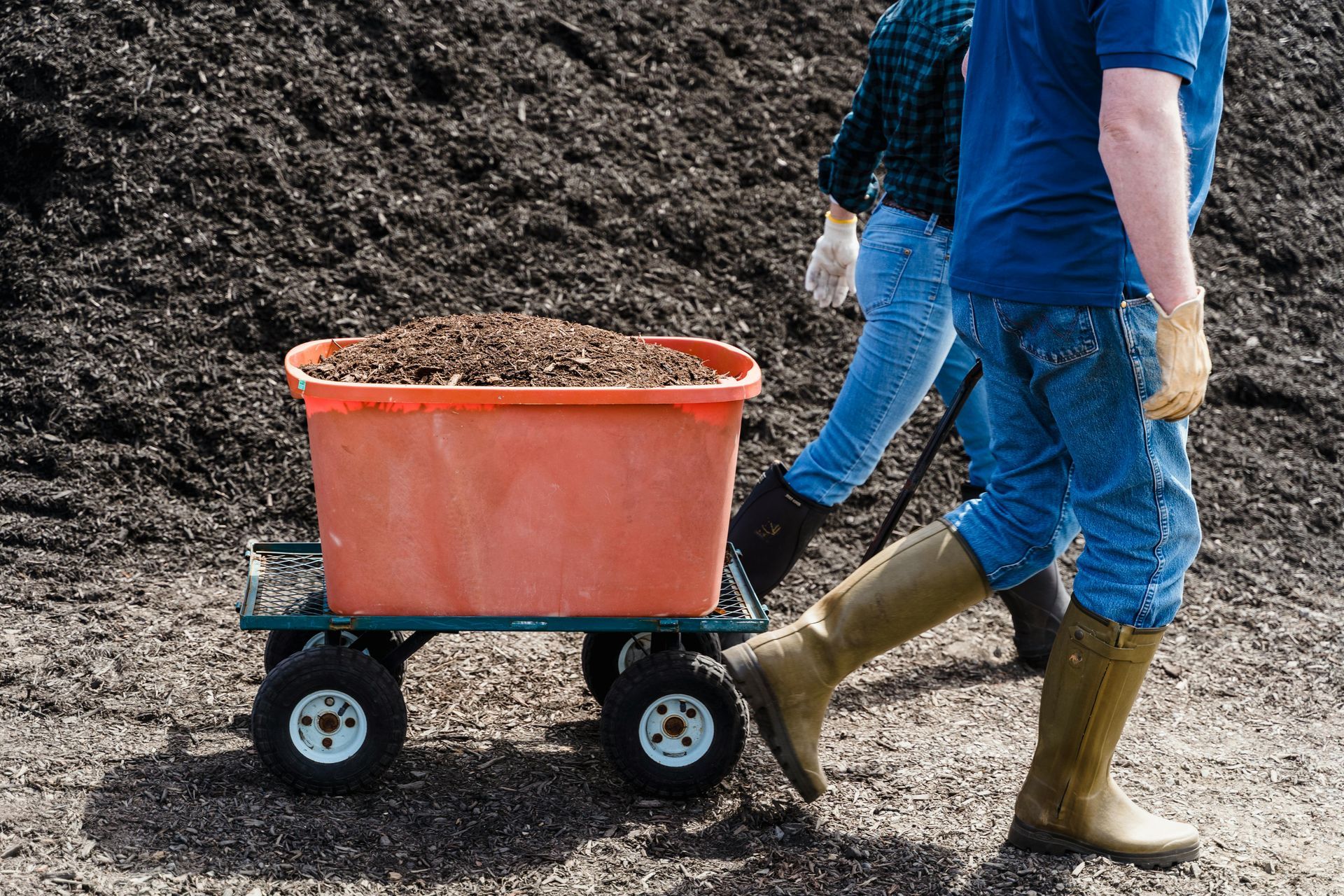 People hauling a red cart full of soil over a dirt pile, wearing blue clothes and boots