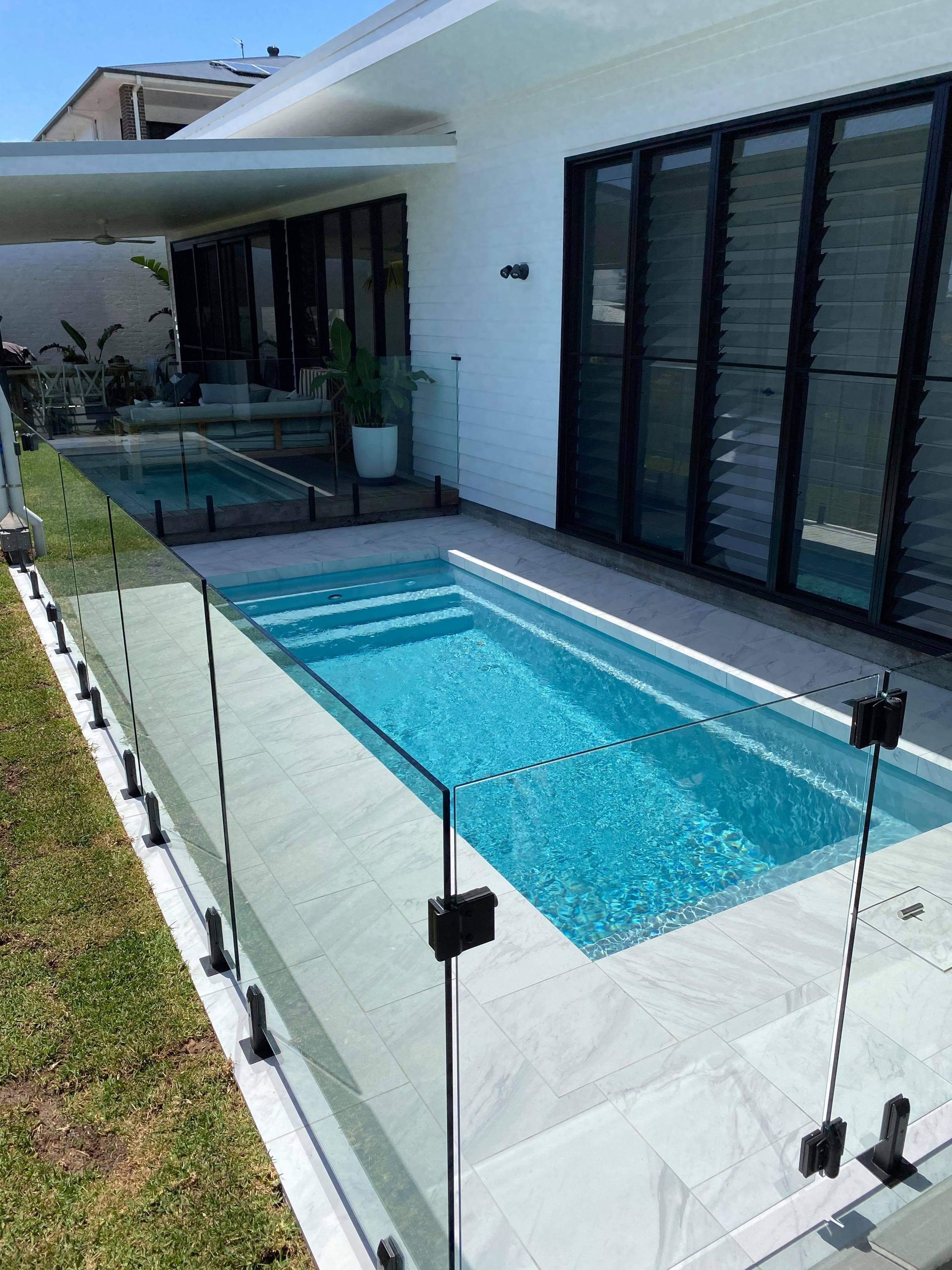 Rectangular pool with turquoise water surrounded by glass fencing and white patio, next to a house — Evoke Pools In Lennox Head, NSW
