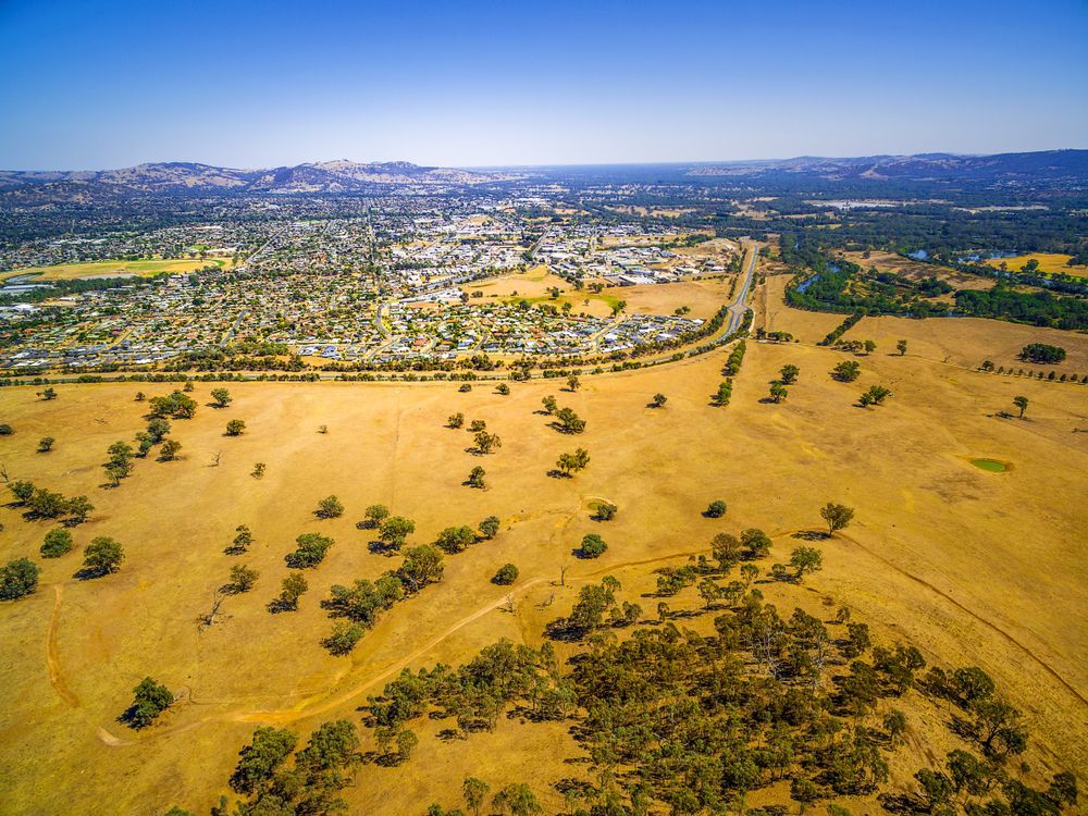 An aerial view of a dry grassy field with trees and a city in the background.