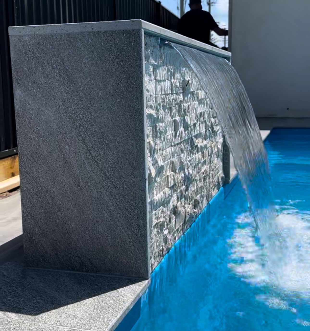 Water feature cascading into a blue pool. Stone and textured wall. Person in background — Evoke Pools In Lennox Head, NSW