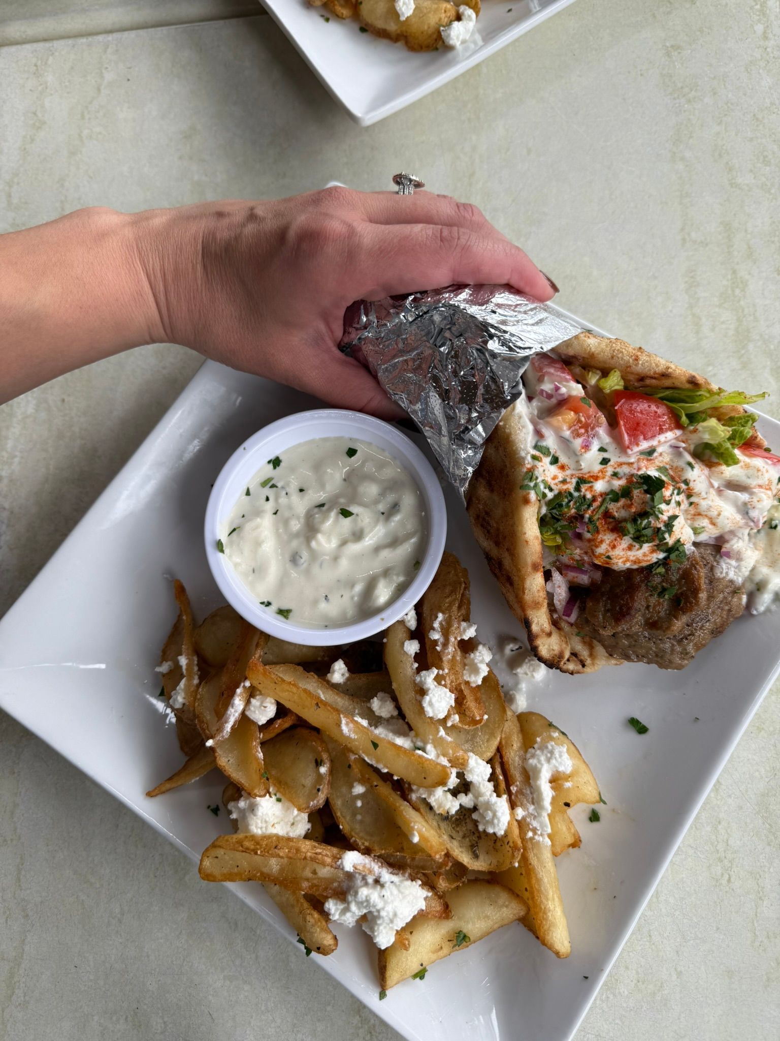 Hand holding a foil-wrapped sandwich with fries and a cup of dipping sauce on a white plate.